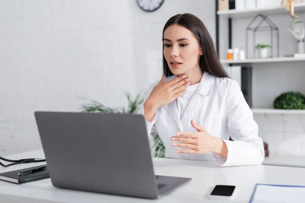 Brunette doctor having video call on laptop and touching neck in clinic — Photo de stock