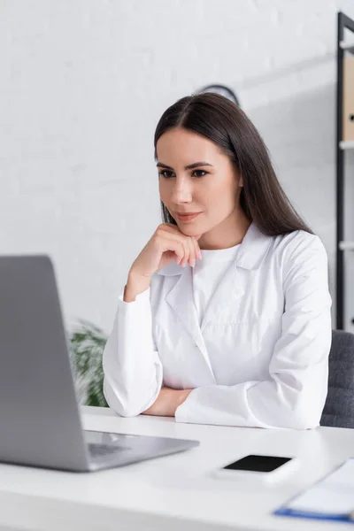 Médico morena en bata blanca mirando borrosa portátil cerca de teléfono inteligente en la clínica - foto de stock