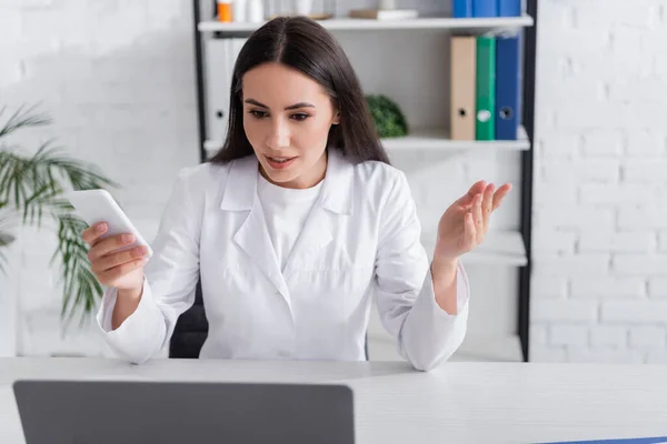 Doctor sosteniendo teléfono inteligente y hablando durante la consulta en línea en el ordenador portátil en la clínica - foto de stock