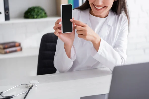 Обрезанный вид улыбающегося врача, указывающего на смартфон во время видеозвонка на ноутбуке в клинике — стоковое фото
