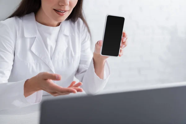 Cropped view of doctor holding cellphone and talking during video call on laptop in clinic - foto de stock