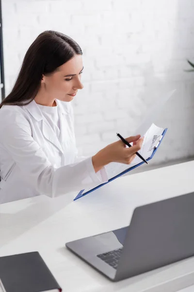 Médico morena sujetando portapapeles cerca del ordenador portátil y portátil en la clínica - foto de stock