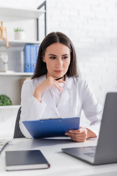 Docteur tenant le stylo et le presse-papiers et regardant l'ordinateur portable flou dans la clinique — Photo de stock