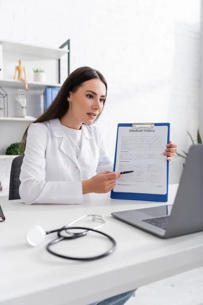 Doctor pointing at medical history on clipboard during online consultation on laptop in clinic — Stock Photo