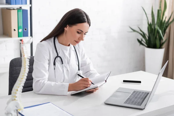 Doctor writing on notebook near devices and blurred spinal model in clinic — Stockfoto