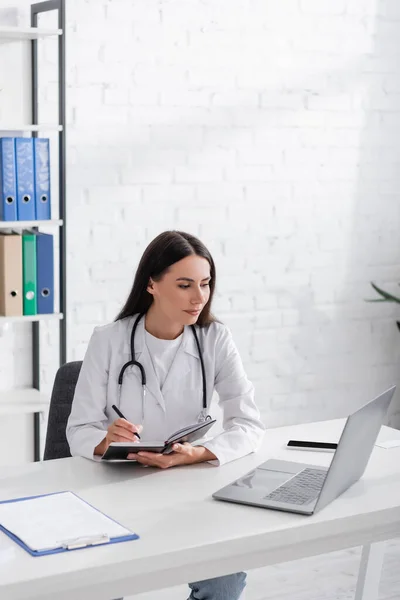 Arzt hält Notizbuch in der Hand und schaut auf Laptop neben Klemmbrett in Klinik — Stockfoto