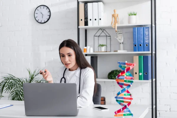 Doctor holding pen while talking during video call on laptop near blurred dna model in clinic — Fotografia de Stock