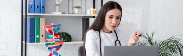 Brunette médecin ayant chat vidéo sur ordinateur portable près de flou modèle ADN à la clinique, bannière — Photo de stock