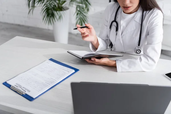 Vista cortada de médico segurando notebook e caneta perto de história médica e dispositivos na clínica — Fotografia de Stock