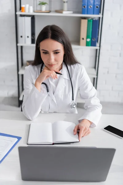 Doctor holding notebook and looking at laptop near clipboard on table in clinic — Stockfoto