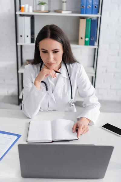 Arzt hält Notizbuch in der Hand und blickt auf Laptop neben Klemmbrett auf Tisch in Klinik — Stockfoto