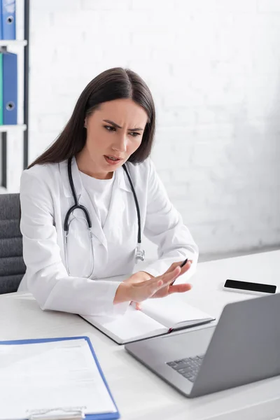 Worried doctor having video call on laptop near clipboard and notebook in clinic — Photo de stock