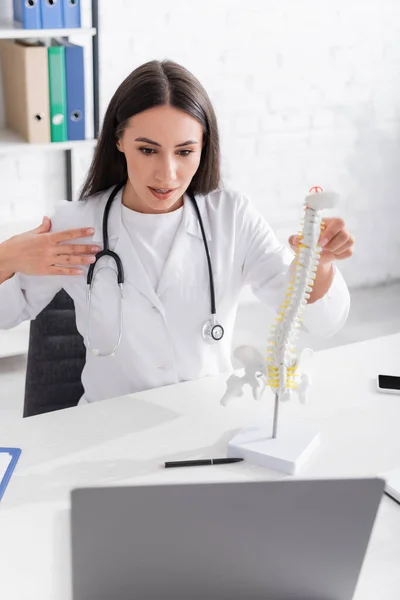 Brunette doctor holding spinal model during online consultation on laptop in hospital — Stock Photo