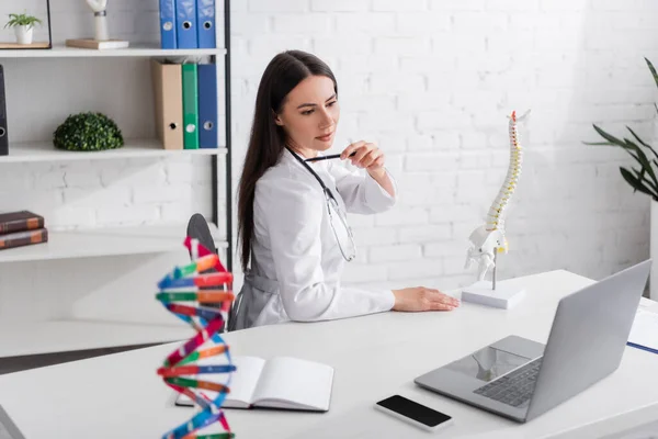 Doctor pointing at shoulder near spinal model during video call on laptop in clinic - foto de stock