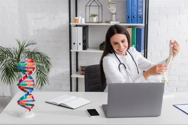 Medico positivo che tiene modello spinale durante la consultazione online su laptop vicino notebook e smartphone in clinica — Foto stock