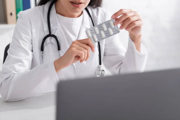 Vista cortada do médico segurando pílulas durante a consulta on-line no laptop na clínica — Fotografia de Stock