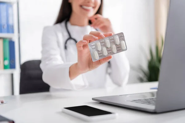 Cropped view of blurred doctor holding blister with pills during video call on laptop in hospital — Photo de stock