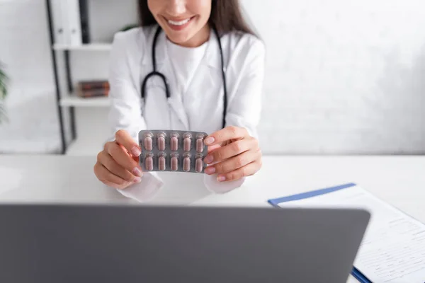 Vista cortada de médico sorrindo segurando blister com pílulas perto de laptop e área de transferência na clínica — Fotografia de Stock