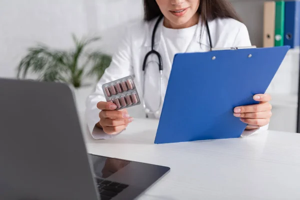 Vista cortada do médico segurando prancheta e pílulas durante a consulta on-line no laptop na clínica — Fotografia de Stock