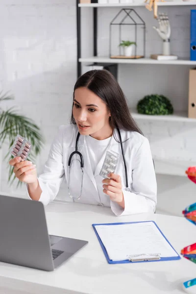 Врач-брюнетка держит таблетки и разговаривает во время видеозвонка на ноутбуке в клинике — стоковое фото