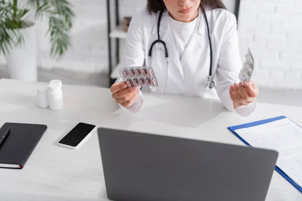Cropped view of doctor holding pills near devices and clipboard on table in clinic — Stock Photo