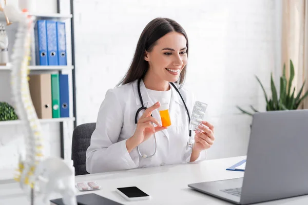 Médico sonriente sosteniendo pastillas y mirando a la computadora portátil durante la videollamada en la clínica - foto de stock