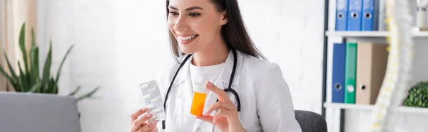 El médico positivo sostiene las píldoras durante la consulta en línea en el ordenador portátil en la clínica, bandera - foto de stock