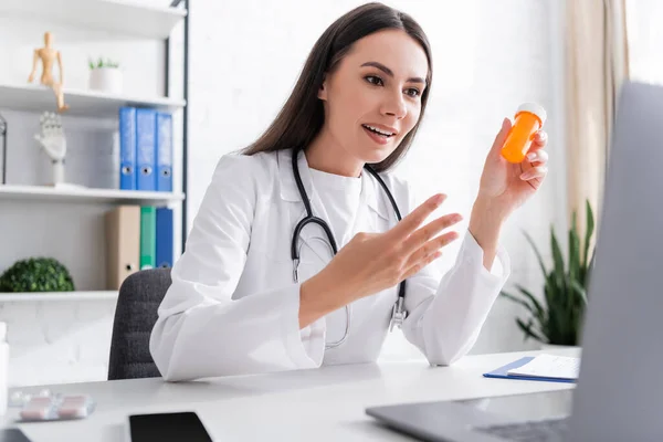 Lächelnder Arzt hält Tabletten in der Hand und gestikuliert bei Videoanruf auf Laptop in Klinik — Stockfoto