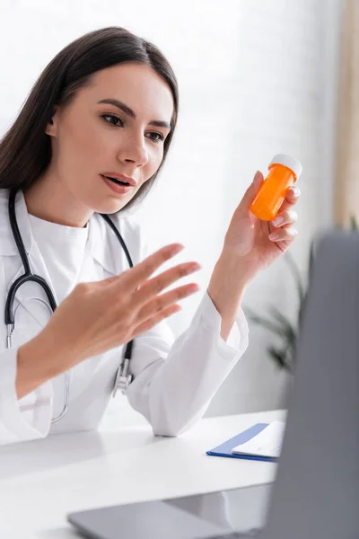 Médico segurando pílulas e gestos durante a consulta on-line no laptop na clínica — Fotografia de Stock