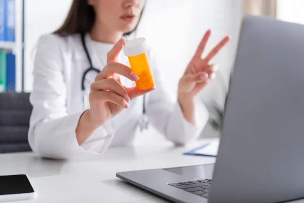 Vista recortada de médico borroso sosteniendo frasco con pastillas cerca de aparatos en la clínica - foto de stock