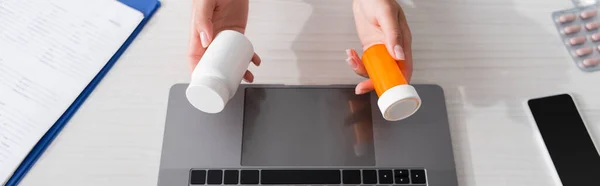 Top view of doctor holding jars with pills near gadgets and clipboard in hospital, banner — Stock Photo