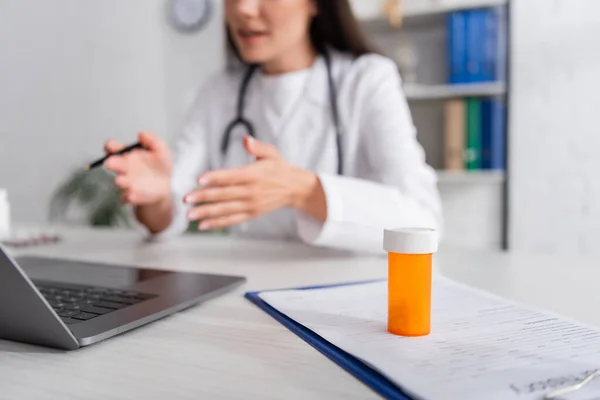 Vue recadrée des pilules sur presse-papiers près du médecin flou ayant appel vidéo sur ordinateur portable dans la clinique — Photo de stock