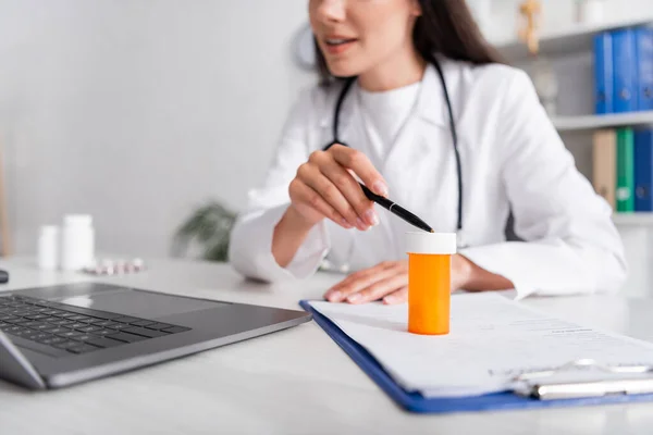 Vista cortada do médico apontando para frasco com pílulas na área de transferência durante a consulta on-line no laptop na clínica — Fotografia de Stock