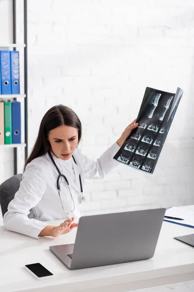 Doctor holding mri scan during online consultation on laptop in clinic — Stock Photo
