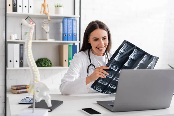 Médecin positif montrant le balayage IRM pendant l'appel vidéo sur ordinateur portable dans la clinique — Photo de stock