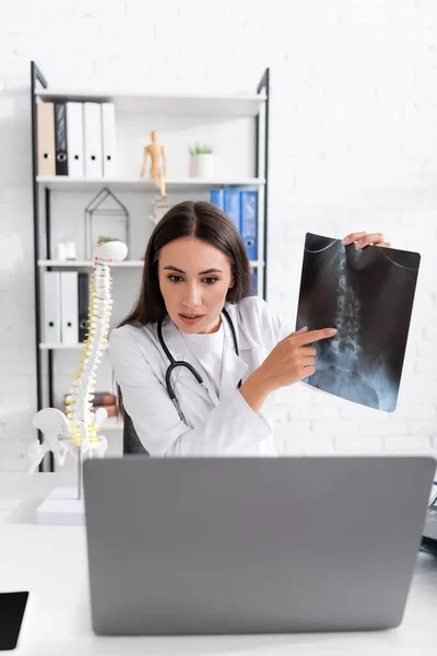 Médecin pointant vers le balayage par rayons X pendant le chat vidéo sur ordinateur portable dans la clinique — Photo de stock