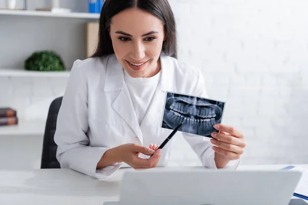 Médico sonriente señalando el escaneo de dientes cerca de la computadora portátil en la clínica - foto de stock