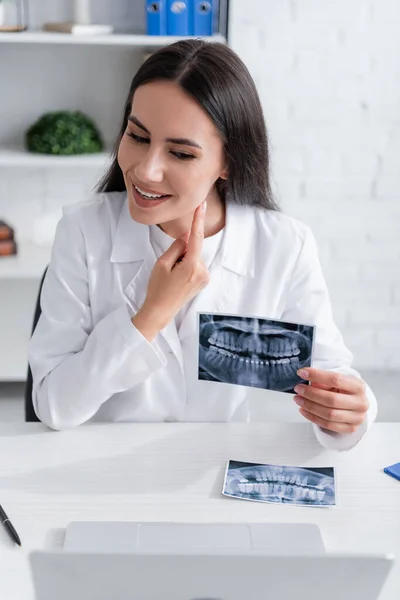 Médico positivo sosteniendo la exploración de los dientes y apuntando a la mejilla durante la videollamada en el ordenador portátil en la clínica - foto de stock