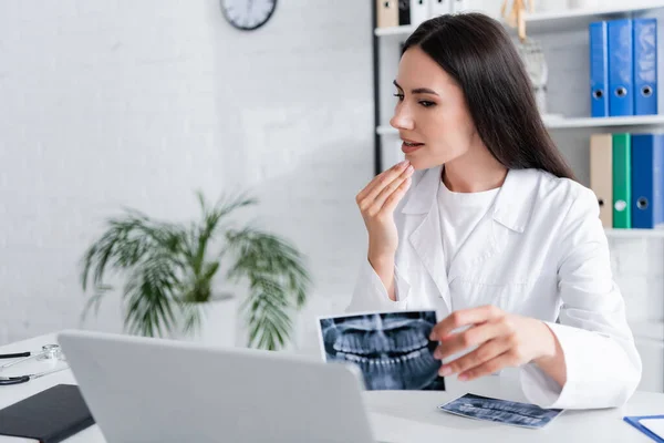 Doctor holding scan of teeth and talking during online consultation on laptop in clinic — Stock Photo