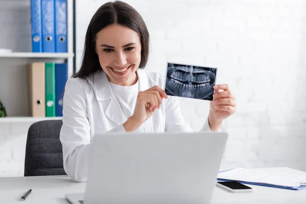 Positive doctor holding scan of teeth during video chat on laptop in hospital — Stock Photo