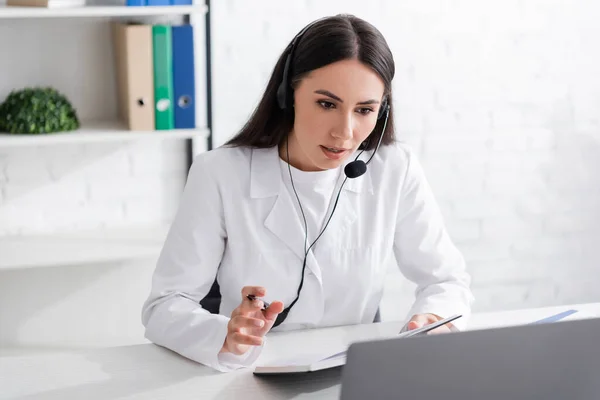Doctor in headset holding notebook during video call on laptop in clinic - foto de stock