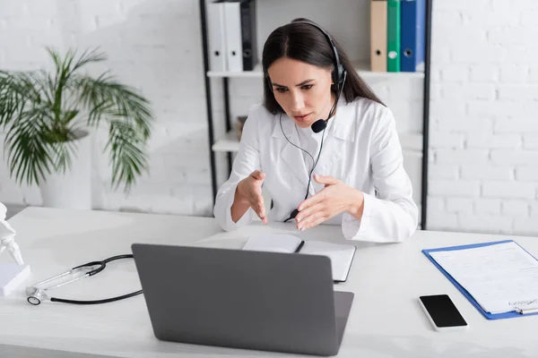 Doutor em fone de ouvido apontando para laptop durante videochamada no laptop no hospital — Fotografia de Stock