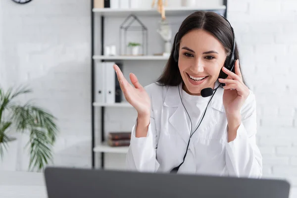 Médico sonriente en auriculares que tiene video consulta en la computadora portátil en la clínica - foto de stock