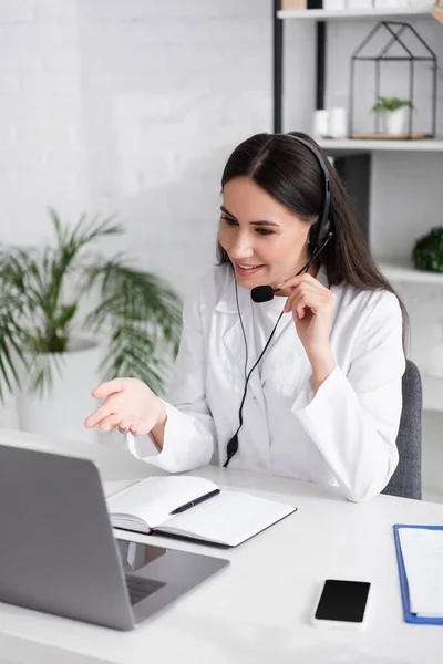 Smiling doctor in headset having video consultation on laptop near notebook and clipboard in clinic — Stock Photo