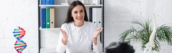 Lächelnder Arzt zeigt mit den Händen, während er in der Nähe der Digitalkamera in der Klinik spricht, Banner — Stockfoto