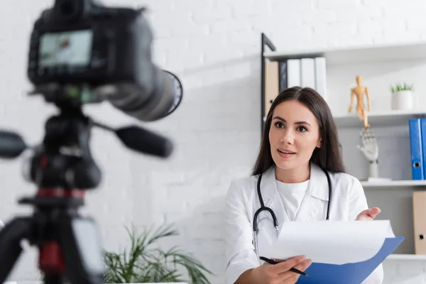 Brunette doctor holding clipboard and pen near blurred digital camera in clinic — Stock Photo