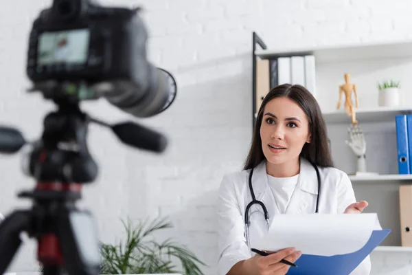 Brünette Ärztin mit Klemmbrett und Stift in der Nähe verschwommener Digitalkamera in Klinik — Stockfoto