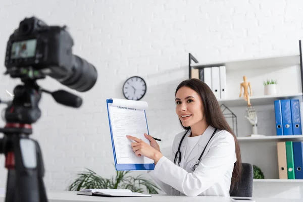 Smiling doctor pointing at clipboard near blurred digital camera in hospital - foto de stock