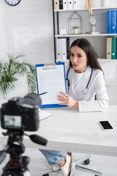 Arzt mit Krankengeschichte nahe verschwommener Digitalkamera in Klinik — Stockfoto