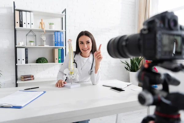 Médico positivo señalando con el dedo cerca del modelo espinal y cámara digital borrosa en la clínica - foto de stock